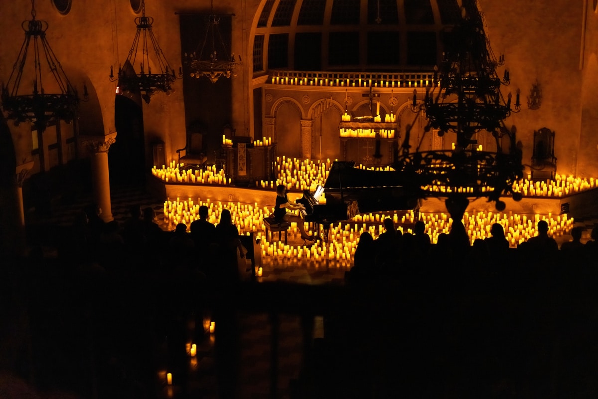A woman is playing the piano at the altar of a church surrounded by hundreds of candles and the silhouette of the audience is visible.
