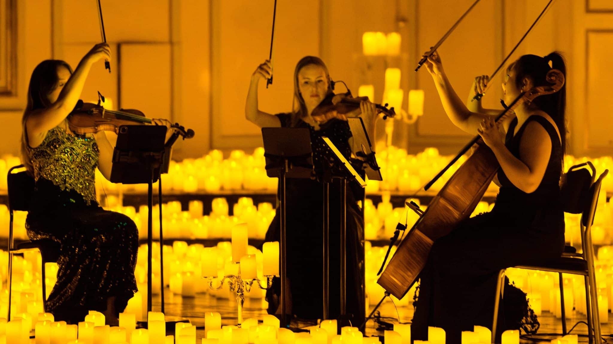 Three musicians play string instruments, surrounded by candles. 